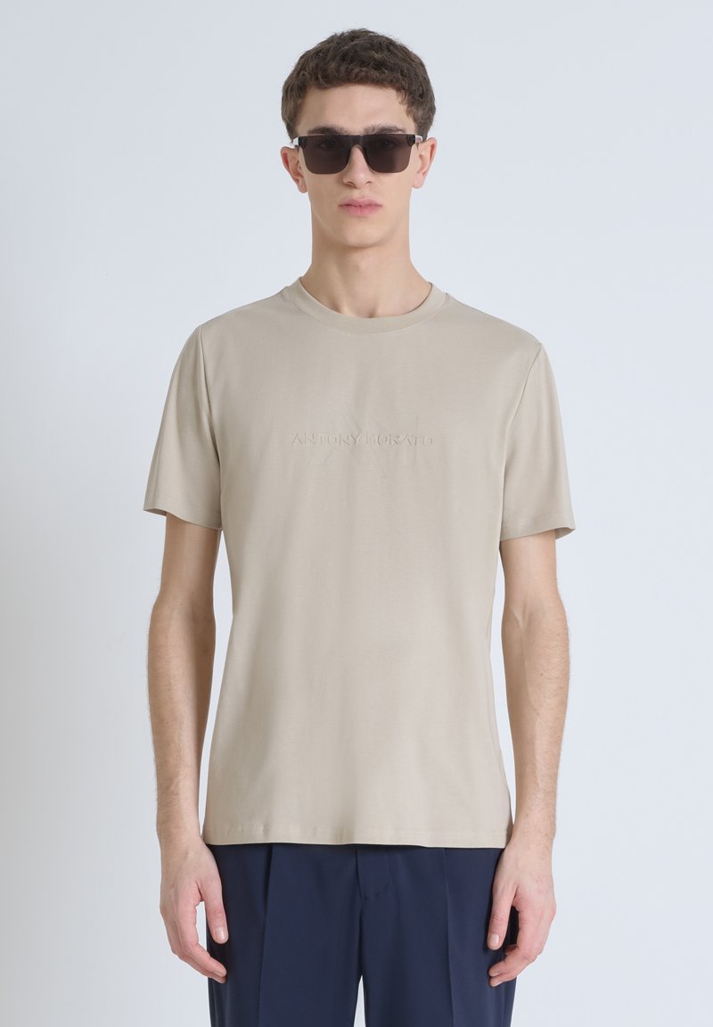 Camiseta de manga corta beige con cuello redondo y detalle de logo en relieve. La tela parece suave y ligera, adecuada para un uso casual.