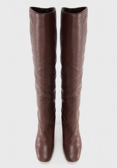 Brązowe, skórzane kozaki sięgające do kolana, o gładkiej fakturze, zaokrąglonym nosku i lekko luźnym kroju. Czysta, minimalistyczna sylwetka.