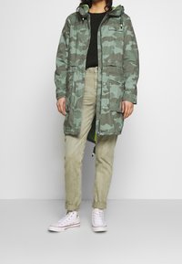 Person wearing a green camouflage hooded jacket, light olive pants rolled at cuffs, black sweater, and white high-top sneakers standing against a plain wall.