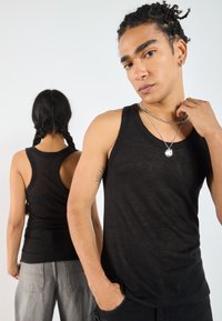 Svart tank top laget av et lett, teksturert stoff med racerback-design. Modell som bærer sølvkjede, med en annen person i bakgrunnen.