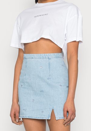 Kobieta w białym skróconym T-shircie z tekstem "MINIMALIST" i jasnoniebieskiej mini spódniczce jeansowej z małymi haftowanymi wzorami i przednim rozcięciem.