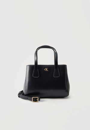 Svart skinnveske med doble håndtak, gullfarget "cK" logo foran, og avtakbar skulderrem med gullspenne på ensfarget bakgrunn.