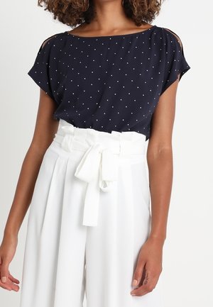 Kobieta ubrana w granatową, krótki rękaw bluzkę w groszki, wpuszczoną w wysokie białe spodnie o szerokich nogawkach z przewiązanym paskiem.