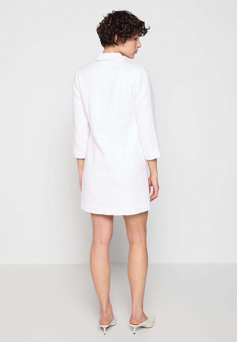 Weißes maßgeschneidertes Kleid mit Kragen, dreiviertel langen Ärmeln und einer strukturierten Oberfläche. Der Saum endet auf Oberschenkelhöhe und wird mit neutralen Absatzschuhen kombiniert.