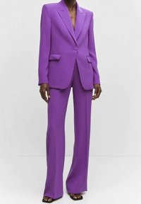 Traje morado a medida con blazer de un solo botón, solapas con muesca y pantalones de pierna recta. La tela tiene una textura suave y un ajuste estructurado.