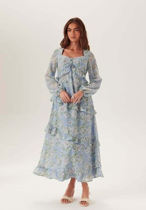 Vrouw met lang bruin haar, gekleed in een blauwe bloemen maxi-jurk met lange mouwen en ruches, staande op blote voeten tegen een effen lichte achtergrond.