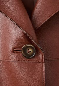 Veste en cuir brun texturé avec un grand bouton marron foncé fixé près du col et des détails de couture visibles.