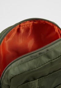 Wnętrze oliwkowej torby materiałowej z jasnopomarańczową podszewką, z zamknięciem na zamek. Zewnętrzna strona ma przeszycia, które dodają faktury.