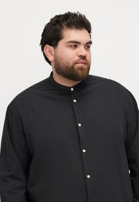Schwarzes Button-Down-Hemd mit Mandarin-Kragen, ausgestattet mit kleinen weißen Knöpfen und einem glatten, strukturierten Stoff. Lässig geschnitten mit langen Ärmeln.
