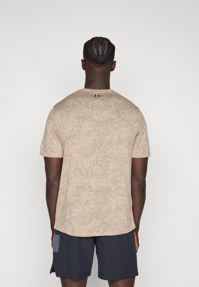 Hombre con cabello trenzado que lleva una camiseta beige de manga corta de Under Armour y pantalones cortos negros, de pie con la espalda hacia el espectador.