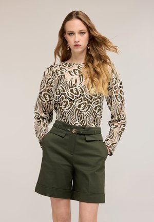 Kobieta z długimi falującymi włosami, ubrana w wzorzystą przezroczystą bluzkę i ciemnozielone, dobrze skrojone, wysokim stanem szorty z paskiem, stoi z rękami w kieszeniach.