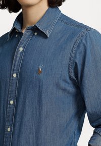 Camisa vaquera en azul medio con cuello abotonado, que presenta un pequeño logo bordado en el pecho izquierdo y botones blancos.