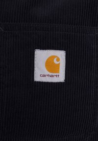 Tessuto in corduroy nero con una toppa bianca quadrata che presenta un logo giallo e la scritta "carhartt" in rosso sul fondo.
