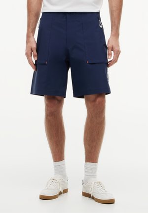 Mężczyzna ubrany w granatowe szorty do kolan z przednimi kieszeniami, białe skarpety i białe trampki, stojący na białym tle.