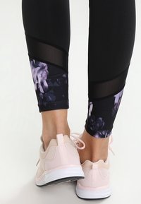 Zwarte legging met bloemenaccenten en meshpanelen, gecombineerd met lichtroze sportschoenen met een textuur bovenkant en een witte zool.
