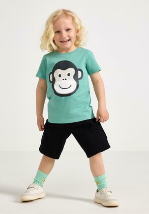 Smilende barn med krøllet blondt hår iført en grønblå trøje med et abeansigt, sorte shorts, grønblå sokker og hvide sneakers.