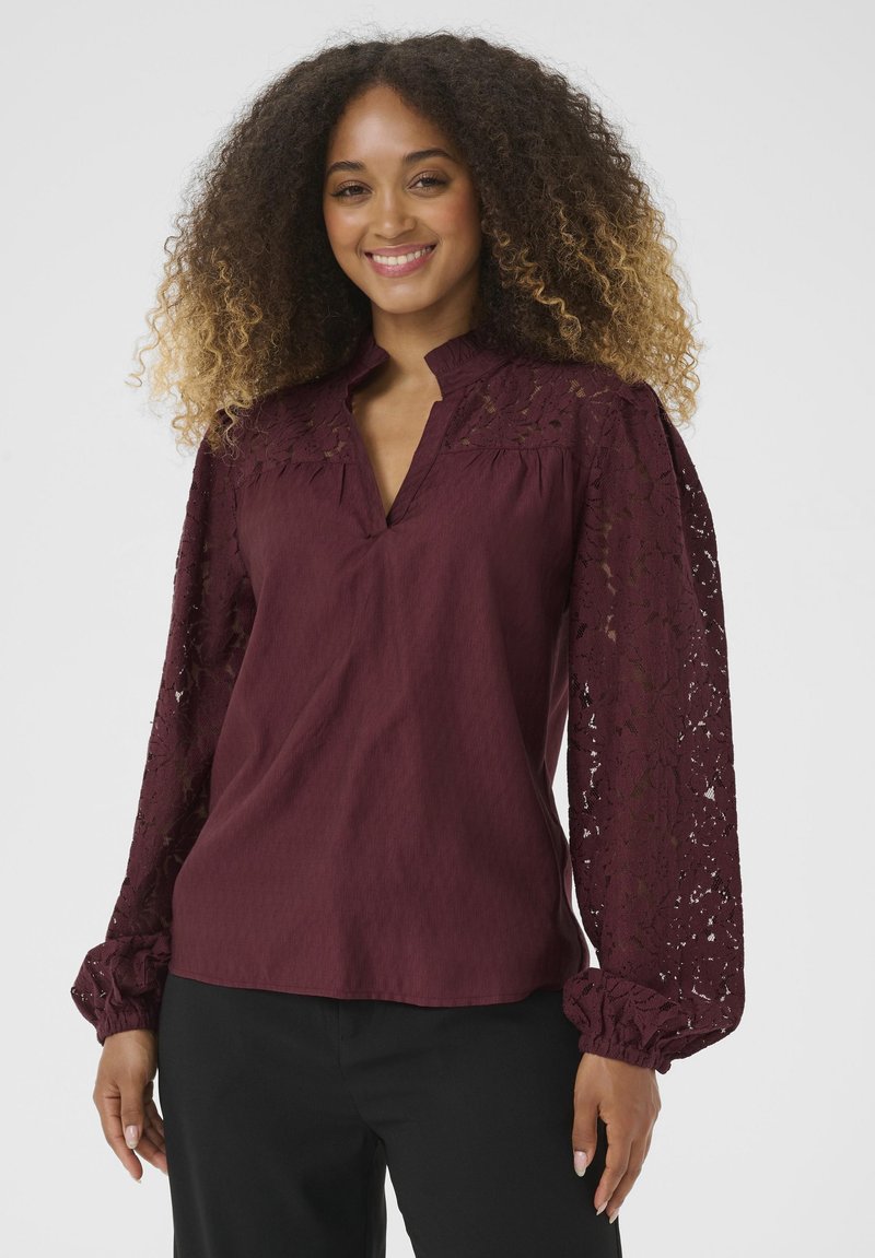 Blouse bordeaux avec un corps texturé subtil, des manches longues en dentelle et un col ouvert. Présente des poignets froncés pour un look volumineux.