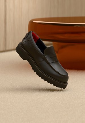 Juodas odinis batų spygliuočio stiliaus batelių pora su raudonu vidiniu pamušalu, ore virš smėlio spalvos kilimo ir šalia medinių baldų.