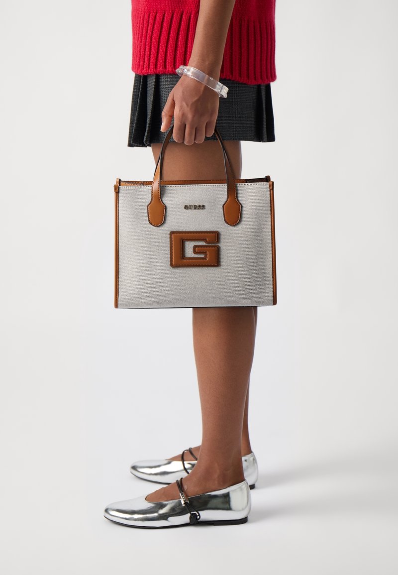 Personne en pull rouge et jupe noire tenant un sac fourre-tout beige et marron avec le logo « G », portant des chaussures plates argentées et un bracelet transparent.