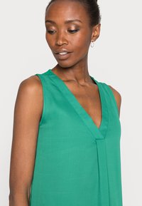 Blusa verde sem mangas com decote em V e design plissado. Feita de tecido suave com um leve brilho. Corte simples e elegante.