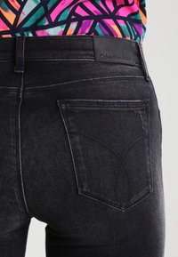 Primer plano del bolsillo trasero de los jeans negros de Calvin Klein con diseño bordado y parte de una blusa de patrón colorido.