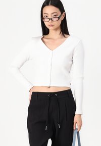 Cardigan corto bianco a coste con scollatura a V e bottoni frontali, abbinato a pantaloni neri a vita alta con coulisse e pince.