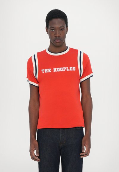 Red short-sleeve t-shirt with contrasting navy stripes on arms and neckline, featuring white "THE KOOPLES" text. Smooth, fitted fabric.