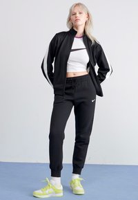 Zwarte trainingsjack met witte zijstrepen, gecombineerd met zwarte joggers met trekkoord. Witte crop top en felgele sneakers met witte accenten.