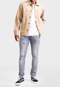 Giacca di jeans beige con bottoni e tasca anteriore, indossata sopra una camicia bianca. Jeans blu chiaro strappati e scarpe da ginnastica nere e bianche.