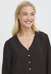 Naeratav naine, kellel on blonde juuksed, kannab musta V-kaelusega pluusi, kus on neli kuldset nööpi, ja vaatab veidi paremale ühtlasel valgel taustal.