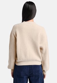 Beige ribbad stickad tröja med rund halsringning och sänkta axlar, med en lös passform. Bärs över mörkblå kordfärgade byxor.