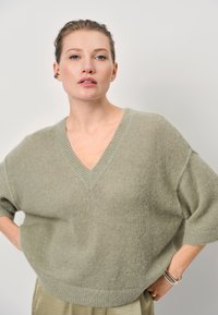 Lichtgroene oversized trui met een V-hals, korte mouwen en een zachte textuur. Beschikt over een geribde zoom en wordt gecombineerd met losse lichte broek.