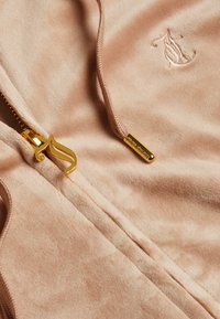Felpa con zip rosa chiaro in tessuto morbido. Presenta dettagli in oro e un logo decorativo dorato. Texture liscia con una leggera lucentezza.