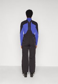 Melni sniega kombinezoni ar regulējamām siksnām, kuri saskaņoti ar zilu un melnu garām piedurknēm kreklu. Audums izskatās ūdensizturīgs ar gludu tekstūru.