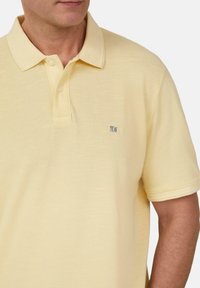 Jasnożółta koszulka polo z teksturowanym materiałem, na trzy guziki, z małą naszywką z logo na lewej piersi. Krótkie rękawy.