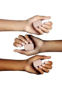 Tre mani tengono bottiglie di smalto per unghie con contenitori in vetro e tappi bianchi. I colori dello smalto variano da tonalità nude chiare a tonalità nude medie.