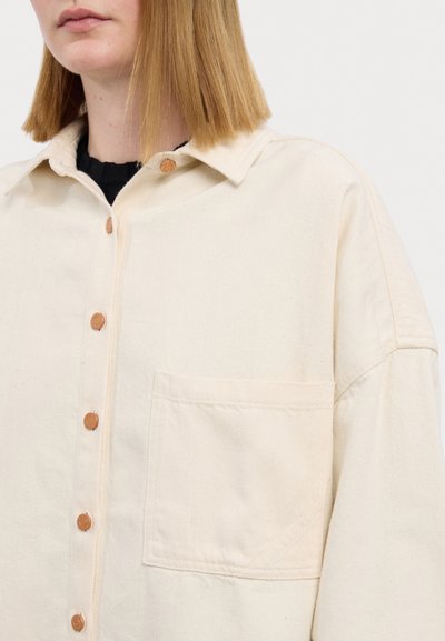 Camisa de botón beige claro hecha de una tela texturizada, con cuello de punta, un bolsillo de parche y acentos de botones marrones.