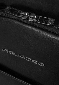 Sac à dos en cuir noir avec une finition texturée, comprenant une fermeture éclair en métal proéminente et un logo embossé « Piquadro » en argent.