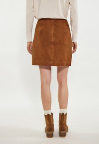Bruine suède mini rok met een gladde textuur, achterkantrits en taille naad. Vergezeld van bruine enkellaarzen met textuurelementen.