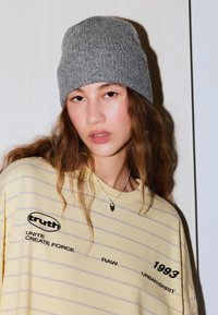 Szürke kötött sapka, sárga csíkos póló fekete szöveggel és grafikákkal, laza, kényelmes illeszkedést és puha anyagot mutatva.