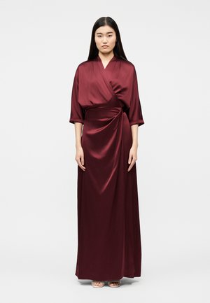 Mujer de pie, vestida con un vestido largo cruzado de satén rojo intenso con mangas tres cuartos y sandalias beige de punta abierta.