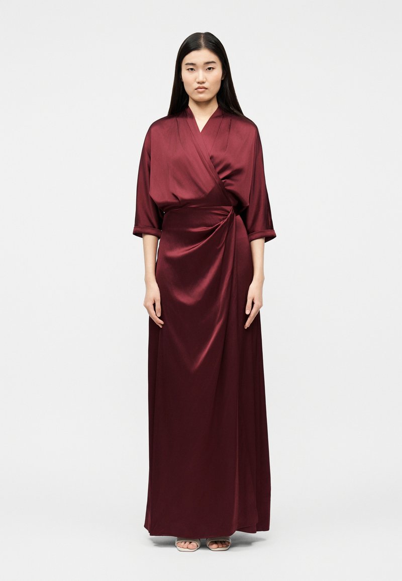 Γυναίκα που στέκεται, φοράει μακρύ, βαθύ κόκκινο σατέν φόρεμα τύπου wrap με μανίκια τριών τετάρτων και μπεζ σανδάλια ανοιχτής μύτης.