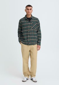 Rutig flannel skjorte i grøn og sort, med knap-lukning, lange ærmer og en brystlomme, parret med beige bukser og sneakers.