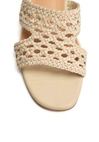 Sandal beige tejido con suela plana, que cuenta con tiras trenzadas cruzadas y un diseño de caja de dedos lisa, fabricado en material sintético.