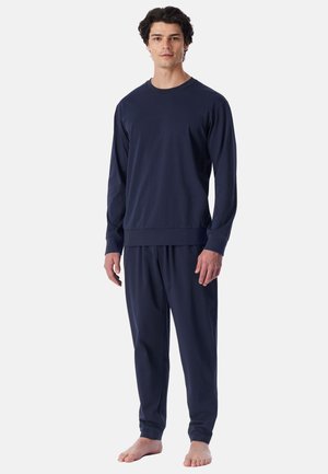 Man staat op blote voeten en draagt een bijpassend donkerblauw shirt met lange mouwen en losse broek tegen een effen lichte achtergrond.