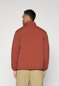 Doudoune rouge à col montant, avec une finition lisse, des poignets élastiques et une coupe décontractée, associée à un pantalon beige.