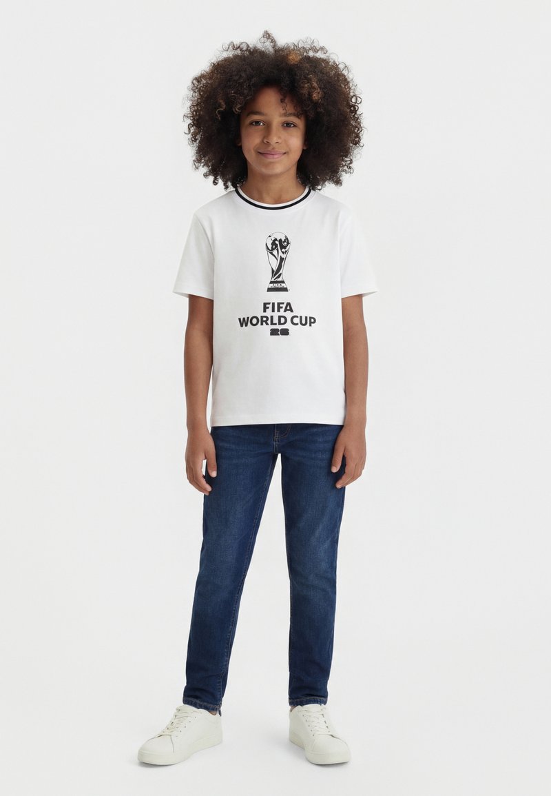 Enfant aux cheveux bouclés portant un t-shirt blanc FIFA World Cup 2022, un jean bleu et des baskets blanches, debout devant un fond uni.