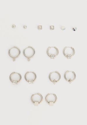 Ensemble de boucles d'oreilles clous en argent et petites boucles d'oreilles créoles avec différents charms géométriques disposés sur un fond blanc.