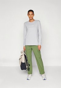 Grå långärmad tröja med Columbia-logotyp, kombinerad med gröna cargobyxor. Modellen håller en ryggsäck och bär vita sneakers med färgglada detaljer.