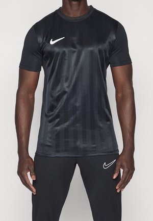 Mężczyzna ubrany w czarną sportową koszulkę i spodnie Nike z białymi logo Nike swoosh, stojący na jednolitym jasnym tle.